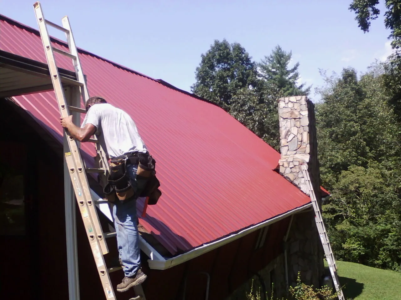 Roofer on a ladder beside a red metal roof and stone chimney — Soffit & Fascia Repair by Roofing Brothers in Midvale